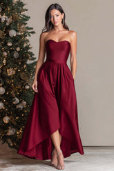 Burgundy satin high-low prom dress with a strapless sweetheart neckline and side pockets, full front view beside a decorated Christmas tree