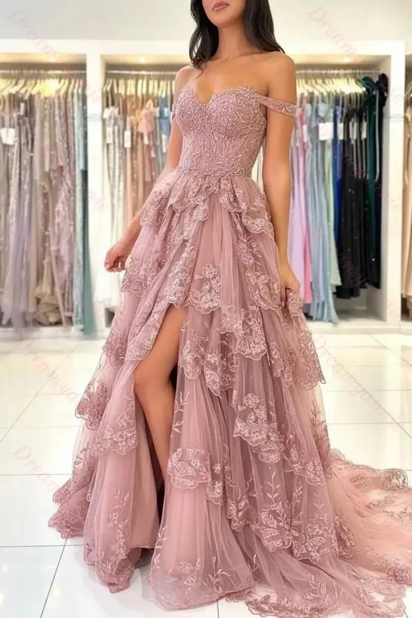 Dusty rose off-the-shoulder bridal dress with tiered lace, tulle layers, and a high leg slit in a boutique setting.